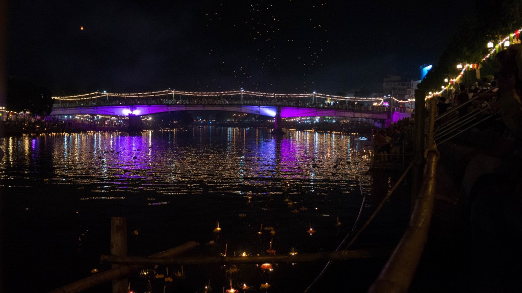 Auf dem Ping River schwimmen leuchtende Boote, während im Hintergrund Himmelslaternen steigen.